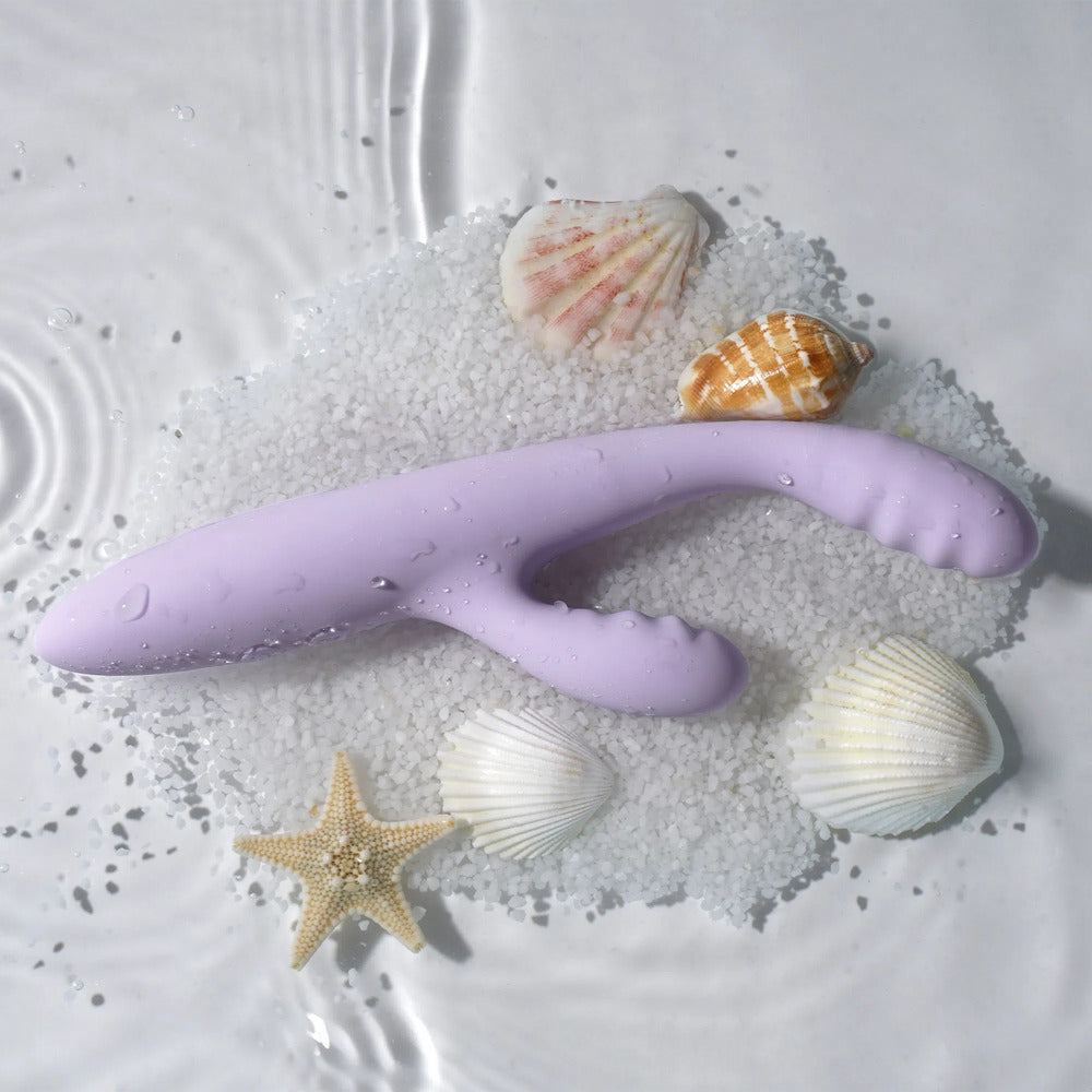 A slim, purple silicone rabbit vibrator with a curved head and a clitoral stimulator, displayed on a bed of白沙 with seashells and a starfish.