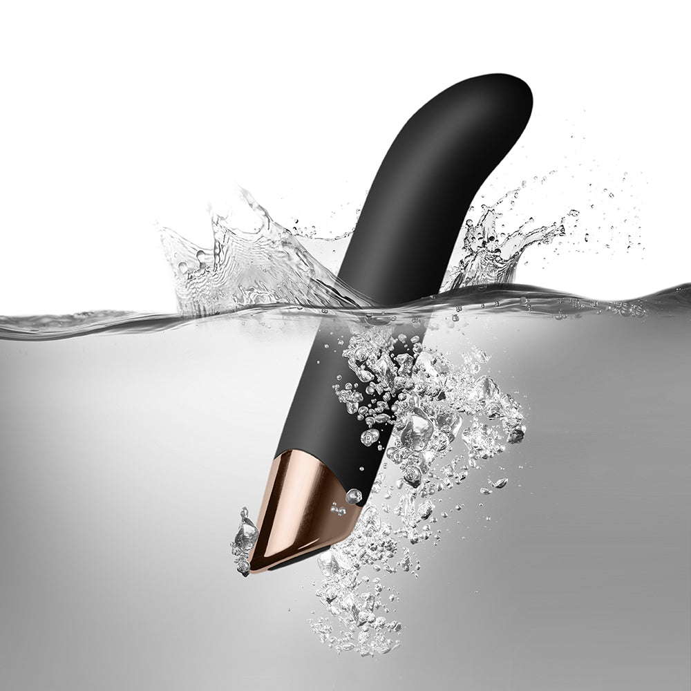 A sleek, black G-spot vibrator with an ergonomic angled tip, resting on a rock, with a branch in the background.
