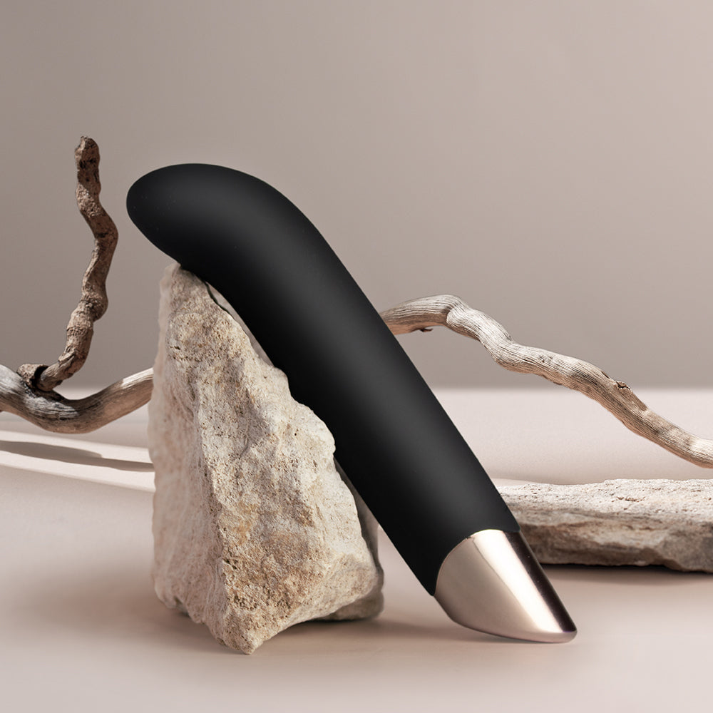 A sleek, black G-spot vibrator with an ergonomic angled tip, resting on a rock, with a branch in the background.