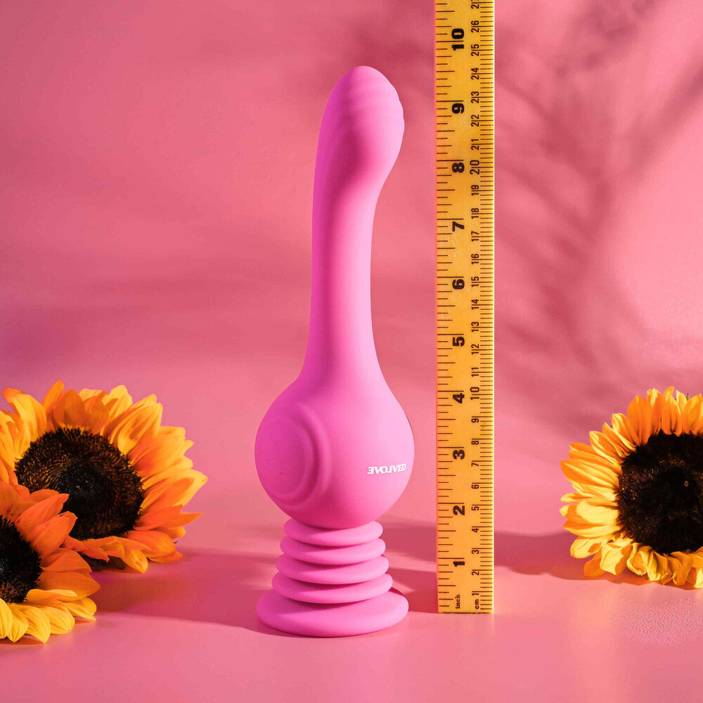 A pink, gyroscopic vibrator with an arched, textured shaft and a accordion-style suction cup base. It stands upright next to a measuring tape, which shows its height, and is accompanied by two sunflowers.