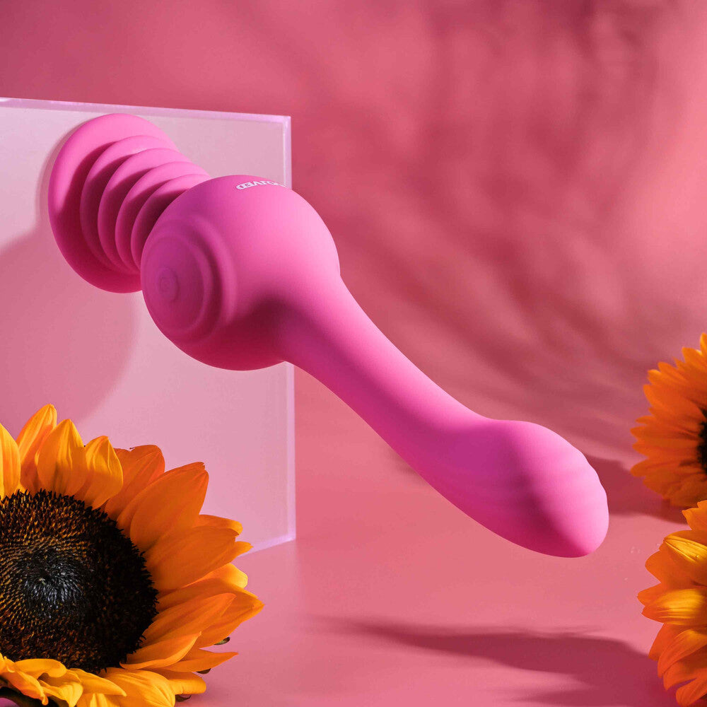 A pink, gyroscopic vibrator with an arched, textured shaft and a accordion-style suction cup base. It stands upright next to a measuring tape, which shows its height, and is accompanied by two sunflowers.
