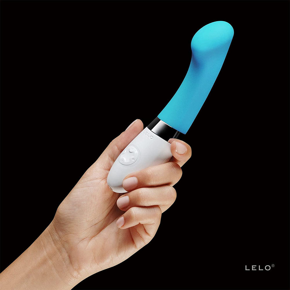 A turquoise blue G-spot vibrator with a sleek, smooth design and a white base. The device has a flattened tip and a curved handle, with a power button and an interface dial on the white stem.