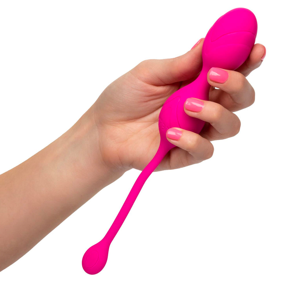 A pink kegel exerciser with a long handle being held in a hand. The device is sleek and has a rounded end, with a small weight attached via a cord.